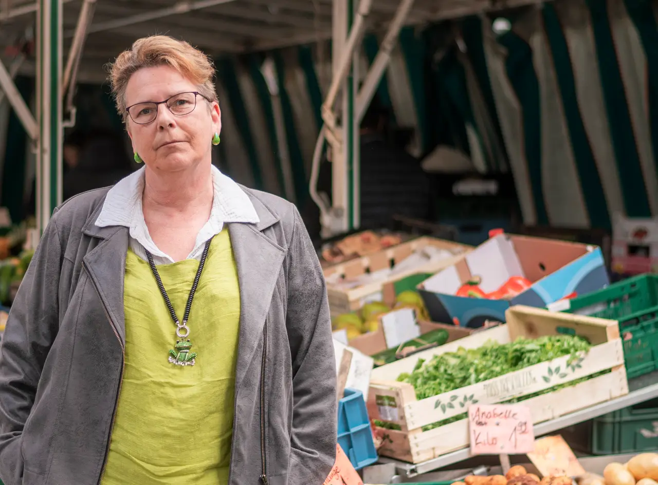 Frau mit Brille und grünem Oberteil steht vor einem Marktstand mit Obst- und Gemüsekisten.